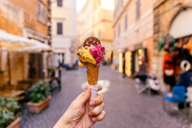 Hand-held gelato cone with three colorful scoops on a sunlit cobblestone street in Italy